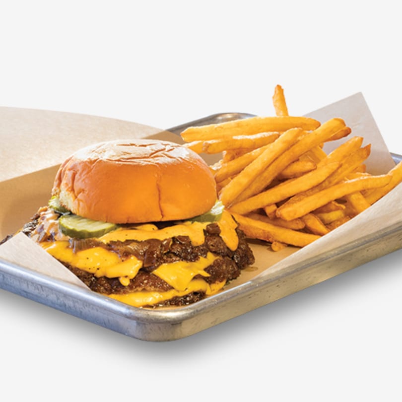 Double cheeseburger with pickles and a side of golden fries on a metal tray.