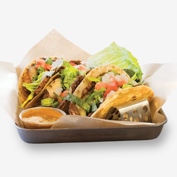 Three tacos with grilled meat, lettuce, tomatoes, and onions in a metal tray with a side of sauce.